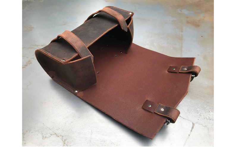 May include: A brown leather motorcycle saddlebag with two straps and buckles. The bag is open and the inside is lined with brown leather.