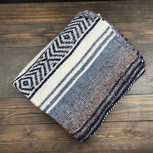 May include: A folded woven blanket featuring a prominent black and white geometric diamond pattern on one side, transitioning into a textured blend of blue, grey, and brown horizontal stripes across the remaining surface of the fabric.