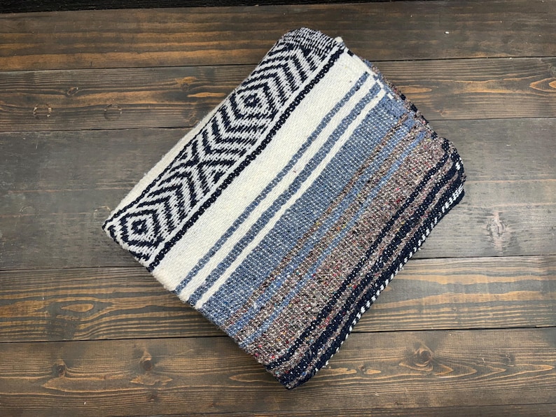 May include: A folded woven blanket featuring a prominent black and white geometric diamond pattern on one side, transitioning into a textured blend of blue, grey, and brown horizontal stripes across the remaining surface of the fabric.