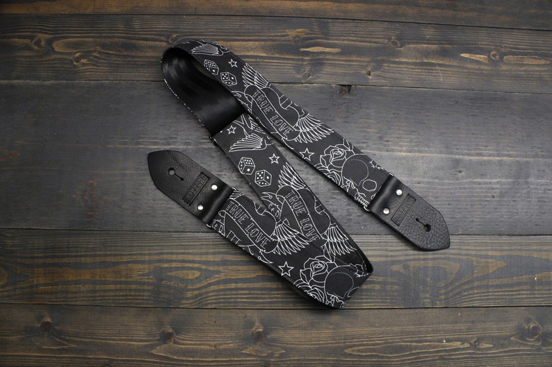 May include: Black guitar strap with white tattoo-style illustrations of roses, dice, stars, and the words "TRUE LOVE." The strap has black leather ends with metal rivets. The strap is lying on a dark wood surface.