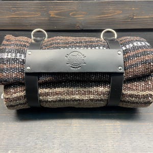 May include: Rolled blanket with a black leather strap and silver rings. The blanket features a woven pattern in shades of brown, black, and white. The leather strap has a stamped emblem. The blanket is rolled up and secured.