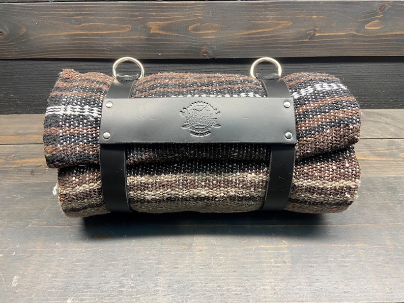 May include: Rolled blanket with a black leather strap and silver rings. The blanket features a woven pattern in shades of brown, black, and white. The leather strap has a stamped emblem. The blanket is rolled up and secured.