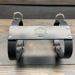May include: Black leather bag carrier with silver metal rings and buckles. The carrier has a stamped logo that reads "Austin, Texas, USA". The carrier is designed to hold a bag or other items.