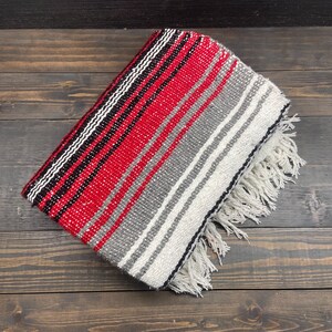 May include: Folded woven blanket with a red, black, white, and gray striped pattern. The blanket has a fringed edge and is displayed on a dark wood surface. The blanket's design is a classic southwestern style.