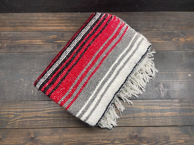 May include: Folded woven blanket with a red, black, white, and gray striped pattern. The blanket has a fringed edge and is displayed on a dark wood surface. The blanket's design is a classic southwestern style.