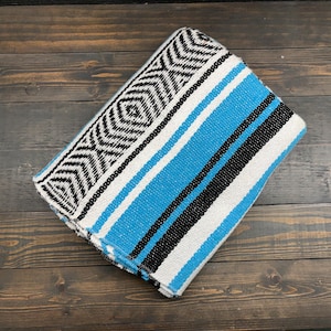 May include: A folded woven blanket featuring a vibrant turquoise blue, black, and white striped pattern. One end of the blanket displays a detailed geometric diamond and chevron design in black and white, contrasting with the solid color stripes.