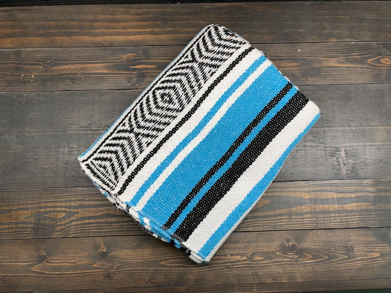 May include: A folded woven blanket featuring a vibrant turquoise blue, black, and white striped pattern. One end of the blanket displays a detailed geometric diamond and chevron design in black and white, contrasting with the solid color stripes.