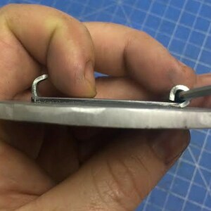 May include: A silver metal belt buckle with a smooth, oval-shaped surface and two metal loops on the back.