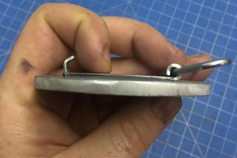 May include: A silver metal belt buckle with a smooth, oval-shaped surface and two metal loops on the back.