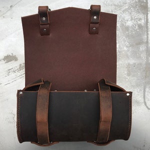May include: A brown leather bag with two straps and a flap. The bag is open and the inside is black.