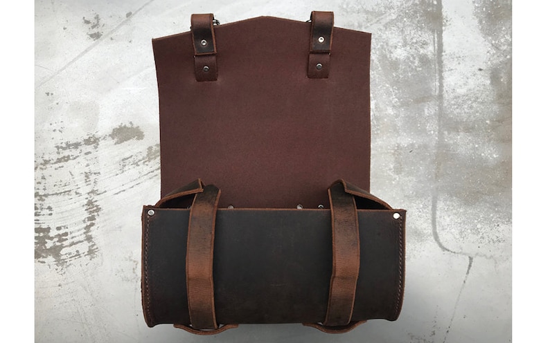 May include: A brown leather bag with two straps and a flap. The bag is open and the inside is black.