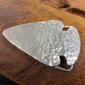 May include: A silver-colored belt buckle with a hammered texture, shaped like an arrowhead. The buckle has a rectangular bar for the belt and is set against a brown leather surface.