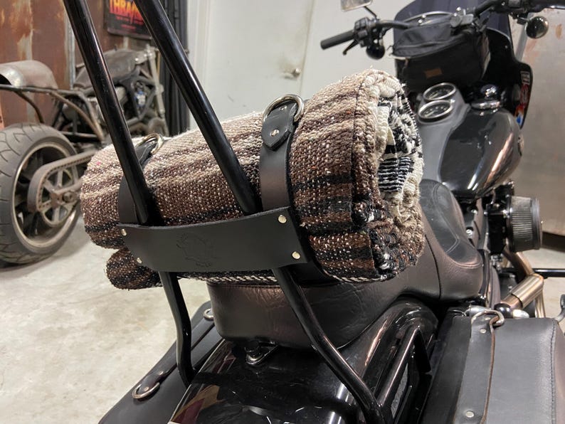 May include: A rolled-up blanket secured to a motorcycle with black leather straps. The blanket features a brown and beige woven pattern. The motorcycle has a black frame and seat.