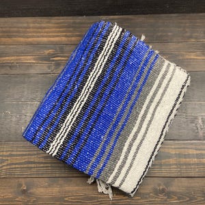May include: A folded blue, black, white, and gray striped blanket with fringe. The blanket is made of woven fabric and is rectangular in shape. The blanket is on a wooden surface.