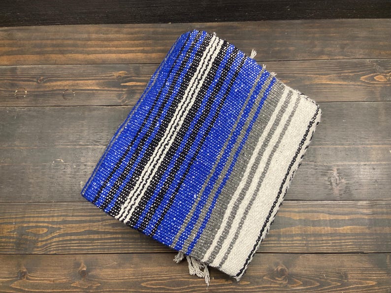May include: A folded blue, black, white, and gray striped blanket with fringe. The blanket is made of woven fabric and is rectangular in shape. The blanket is on a wooden surface.