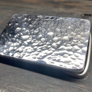 Puede incluir: Hebilla de cinturón rectangular de metal martillado con superficie texturizada. La hebilla es de color plateado y tiene un diseño simple y clásico. La hebilla se muestra sobre una superficie de madera oscura.