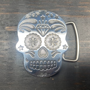 Pode incluir: Uma fivela de cinto de metal prateado em forma de caveira de açúcar com detalhes intrincados, incluindo um diamante, flores e estrelas.