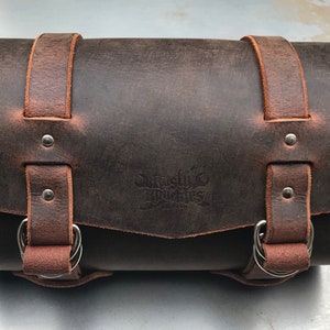 May include: A brown leather motorcycle saddlebag with two buckles and a metal ring on each side. The bag has a distressed look and a logo embossed on the front that says "Rustic Branches".