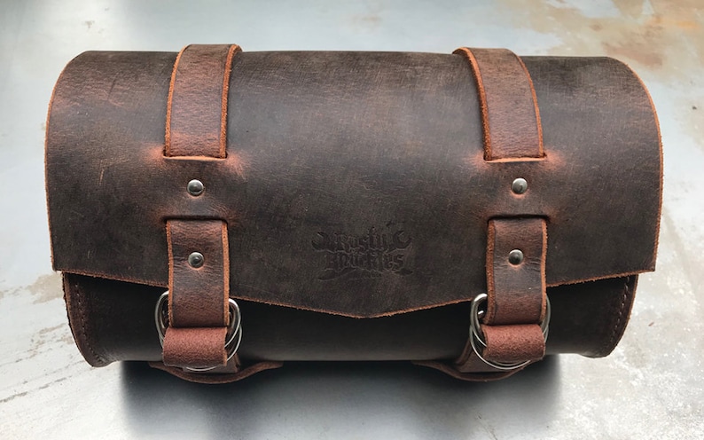 May include: A brown leather motorcycle saddlebag with two buckles and a metal ring on each side. The bag has a distressed look and a logo embossed on the front that says "Rustic Branches".