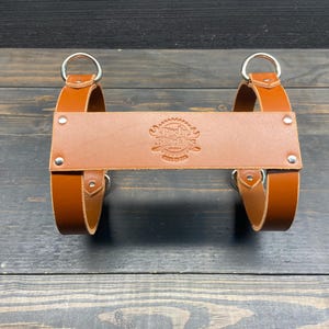May include: A brown leather product with two silver rings and a central leather piece with an embossed design. The product has a rustic appearance and is likely a carrying accessory.