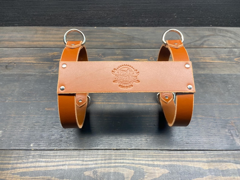 May include: A brown leather product with two silver rings and a central leather piece with an embossed design. The product has a rustic appearance and is likely a carrying accessory.