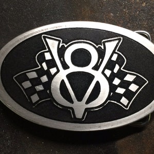 Könnte beinhalten: Eine ovale Gürtelschnalle aus Silber mit schwarzem Hintergrund. Die Schnalle zeigt ein stilisiertes "V8"-Logo mit karierten Flaggen auf beiden Seiten.