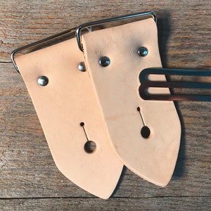 May include: Two light brown leather straps with silver buckles and a silver keeper. The straps have a pointed end and a hole for the buckle.