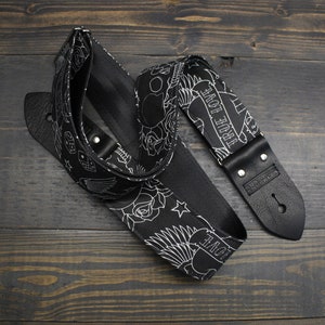 May include: Black guitar strap with a white tattoo-style design featuring roses, stars, dice, and the words "TRUE LOVE." The strap has black leather ends and metal rivets. The strap is lying on a dark wooden surface.