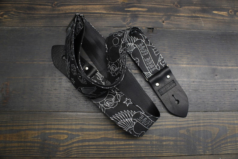 May include: Black guitar strap with a white tattoo-style design featuring roses, stars, dice, and the words "TRUE LOVE." The strap has black leather ends and metal rivets. The strap is lying on a dark wooden surface.