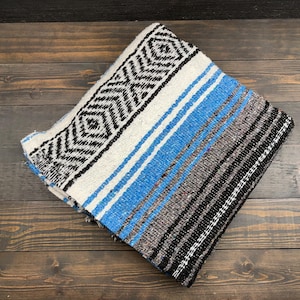 May include: A folded woven blanket featuring a prominent black and white diamond geometric pattern on one side, transitioning into horizontal stripes of bright blue, grey, and black. The textile has a textured appearance with visible weave.