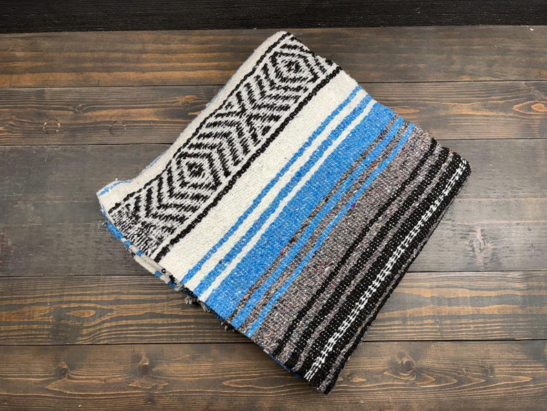 May include: A folded woven blanket featuring a prominent black and white diamond geometric pattern on one side, transitioning into horizontal stripes of bright blue, grey, and black. The textile has a textured appearance with visible weave.