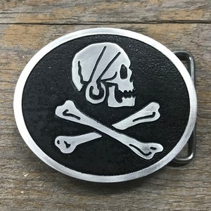 May include: A silver belt buckle with a black background and a silver skull and crossbones design. The skull is wearing a bandana.