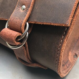 May include: Close-up of a brown leather bag with a silver buckle and strap. The bag has a stitched seam and is made of a thick, durable leather.