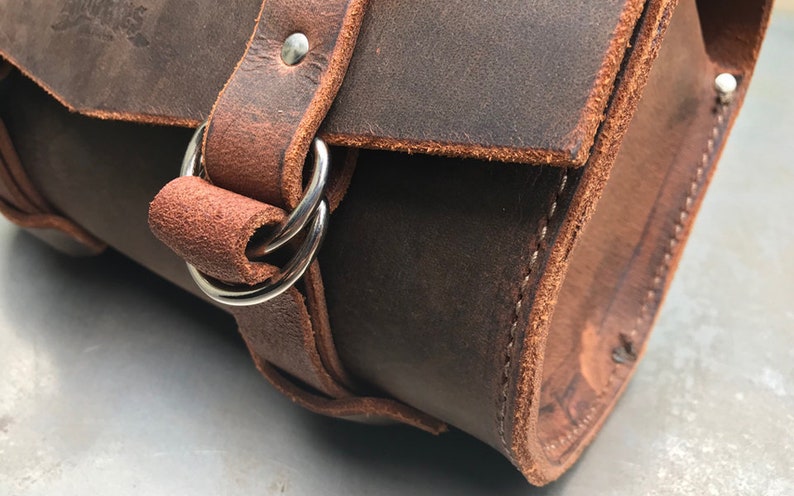 May include: Close-up of a brown leather bag with a silver buckle and strap. The bag has a stitched seam and is made of a thick, durable leather.