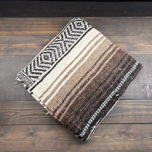 May include: A folded woven blanket featuring a geometric diamond pattern in black and white on one side, transitioning into a series of horizontal stripes in shades of cream, tan, light brown, and dark brown across the rest of the fabric surface.