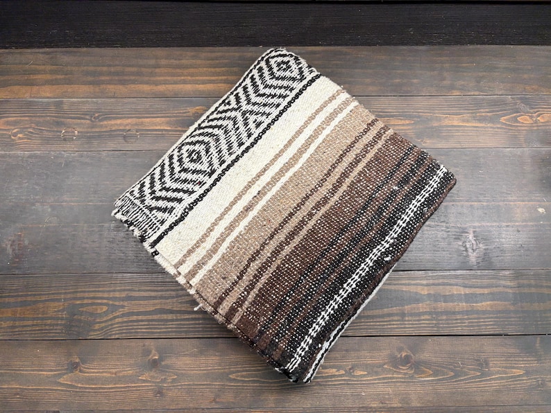 May include: A folded woven blanket featuring a geometric diamond pattern in black and white on one side, transitioning into a series of horizontal stripes in shades of cream, tan, light brown, and dark brown across the rest of the fabric surface.