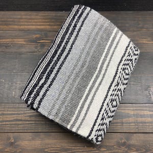 May include: Folded woven blanket with a striped pattern in shades of gray, black, and white. The blanket features a geometric design on one end and is made of a soft, textured material. The blanket is folded neatly on a wooden surface.