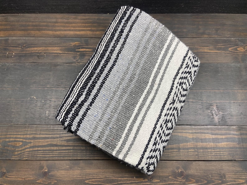 May include: Folded woven blanket with a striped pattern in shades of gray, black, and white. The blanket features a geometric design on one end and is made of a soft, textured material. The blanket is folded neatly on a wooden surface.