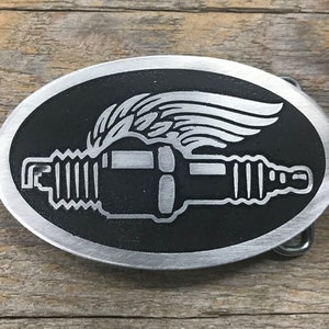 May include: A silver oval belt buckle with a black background. The buckle features a stylized image of a spark plug with a wing behind it.