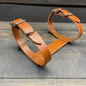 May include: Two brown leather dog collars with silver-colored metal hardware. The collars are connected by a flat leather strap. The collars have a buckle and a metal ring for leash attachment. The background is a dark wooden surface.