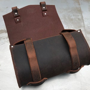 May include: A brown leather saddlebag with two straps and buckles. The bag is open and the inside is lined with a burgundy fabric.