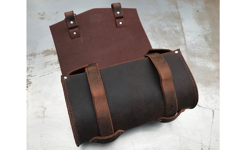 May include: A brown leather saddlebag with two straps and buckles. The bag is open and the inside is lined with a burgundy fabric.