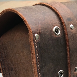 May include: Close-up of a brown leather bag with silver rivets and stitching. The bag has a flap closure and a curved shape.