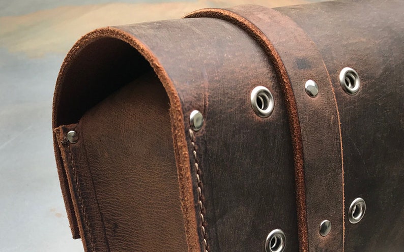 May include: Close-up of a brown leather bag with silver rivets and stitching. The bag has a flap closure and a curved shape.