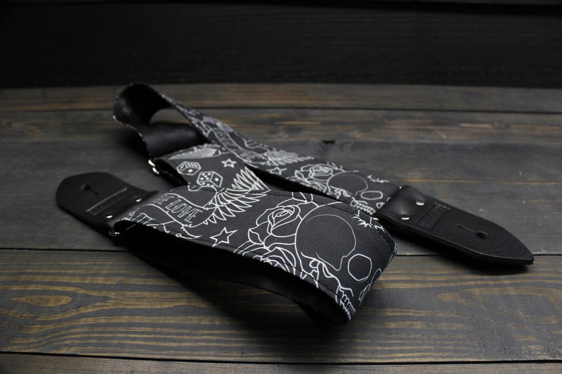 May include: Black guitar strap with a white tattoo-style design featuring skulls, roses, dice, and the word "LOVE". The strap is made of a durable material and has black leather ends.