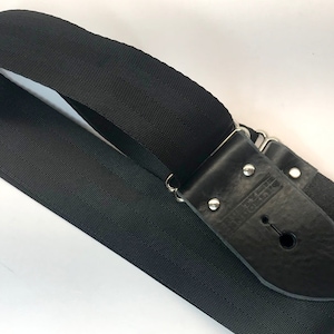 Może przedstawiać: Czarny pasek do gitary ze skórzanymi końcówkami i srebrnym osprzętem. Pasek wykonany jest z tkaniny tkanej i ma wygrawerowane słowo "Leather" na skórzanym końcu.