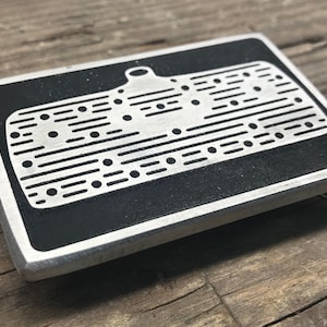 Puede incluir: Una hebilla de cinturón de metal plateado con un diseño de esmalte negro de un desempañador de la ventana trasera de un automóvil. El desempañador tiene una serie de líneas y puntos.