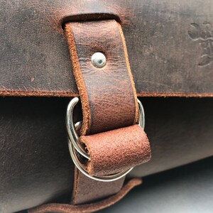 May include: Close-up of a brown leather bag with a silver buckle and two rings. The bag has a stitched edge and the text "Domestic Fittings" is embossed on the top flap.
