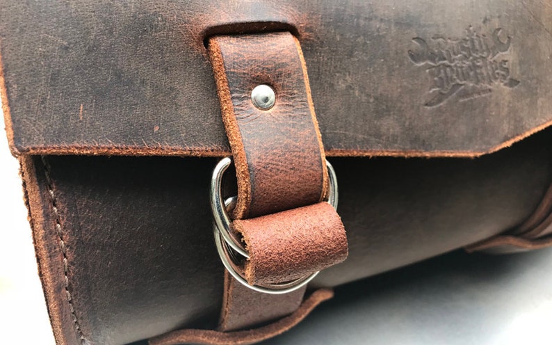May include: Close-up of a brown leather bag with a silver buckle and two rings. The bag has a stitched edge and the text "Domestic Fittings" is embossed on the top flap.
