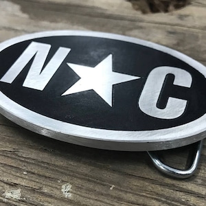 May include: A silver oval belt buckle with a black background and a white star and the letters "N*C" in white.
